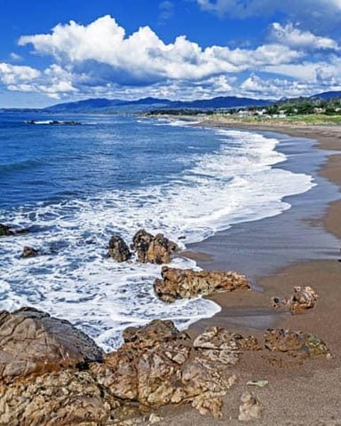Sandy beach with rocky outcrops, white-capped waves, coastal town and mountains under blue sky