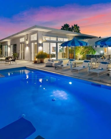 Modern house with illuminated pool, blue umbrellas, lounge chairs, and palm trees at sunset