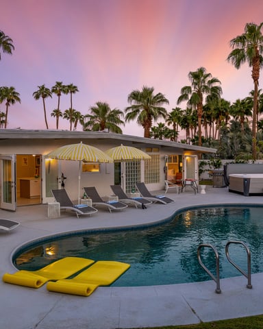 Backyard pool at dusk with lounge chairs, yellow umbrellas, hot tub, and mid-century modern house surrounded by palm trees at sunset.
