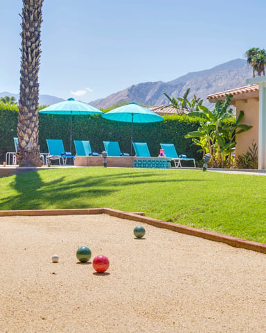 Bocce ball court with colored balls in foreground, lawn with lounge chairs and turquoise umbrellas, palm trees, and mountain views beyond.