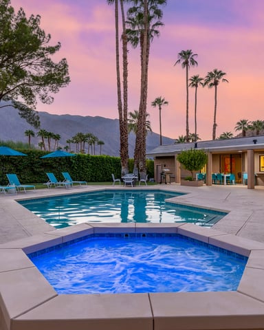 Modern home with pool and spa at sunset, palm trees and mountains in background, blue lounge chairs and umbrellas