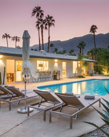 Modern single-story home with pool, lounge chairs, and umbrellas at dusk, palm trees and mountains in background