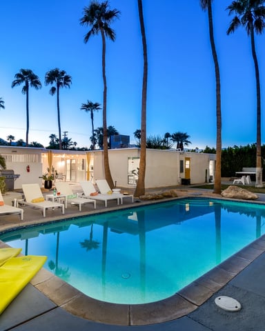 Mid-century modern house with turquoise pool, lounge chairs, and tall palm trees at dusk