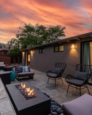 Modern patio at dusk with fire pit, lounge chairs, and illuminated house against dramatic sunset sky and mountains