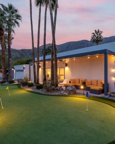 Modern white house with illuminated patio, tan seating, putting green, tall palms, and mountain views at dusk