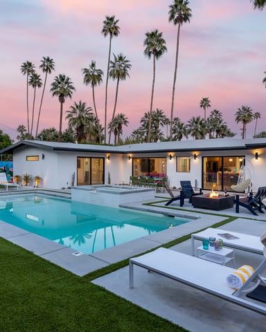 Modern white house with pool, lounge chairs, fire pit, and palm trees at dusk