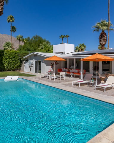 Modern mid-century home with swimming pool, orange umbrellas, lounge chairs, and palm trees against desert mountain backdrop