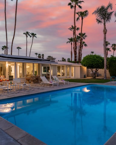 Modern house with illuminated interior, blue swimming pool, lounge chairs, and palm trees at sunset