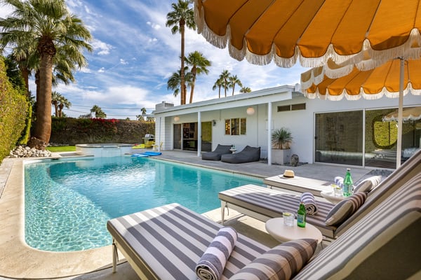 Modern white house with turquoise pool, striped lounge chairs, orange umbrellas, and palm trees in desert landscape