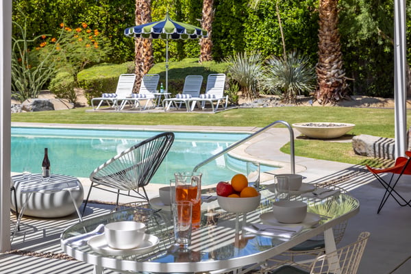 Backyard pool area with patio table, lounge chairs, striped umbrella, and manicured lawn with palm trees
