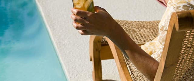 Person relaxing in lounge chair by pool holding glass of iced beverage