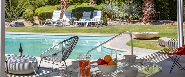 Backyard pool area with patio table, lounge chairs, striped umbrella, and manicured lawn with palm trees